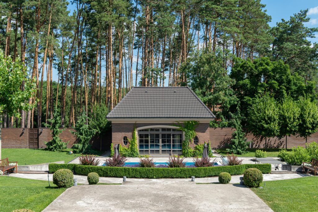 A modern shed overlooks a luxurious patio with a swimming pool, surrounded by lush greenery and tall trees.